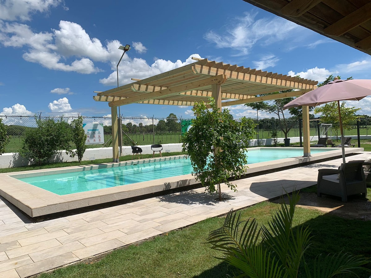 A large outdoor area features a private swimming pool surrounded by a wooden pergola. The pool is framed by a spacious patio, with sun loungers and greenery nearby. The background includes a sports court and a clear blue sky with scattered clouds.