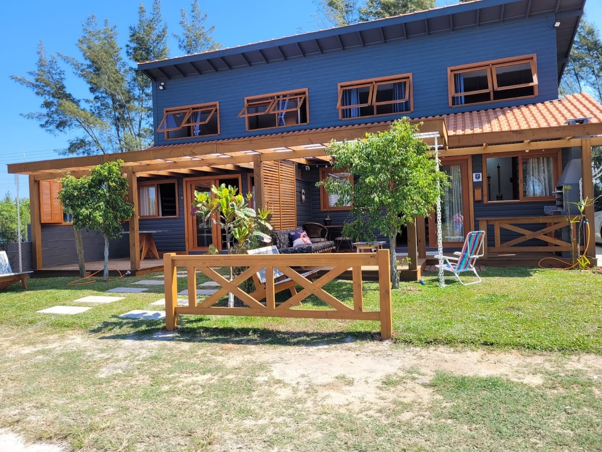 A two-story house with a wooden exterior is surrounded by greenery. The front porch features comfortable seating and is adorned with plants. Large windows provide natural light, while neatly arranged stepping stones lead through the yard, indicating a well-maintained outdoor space.