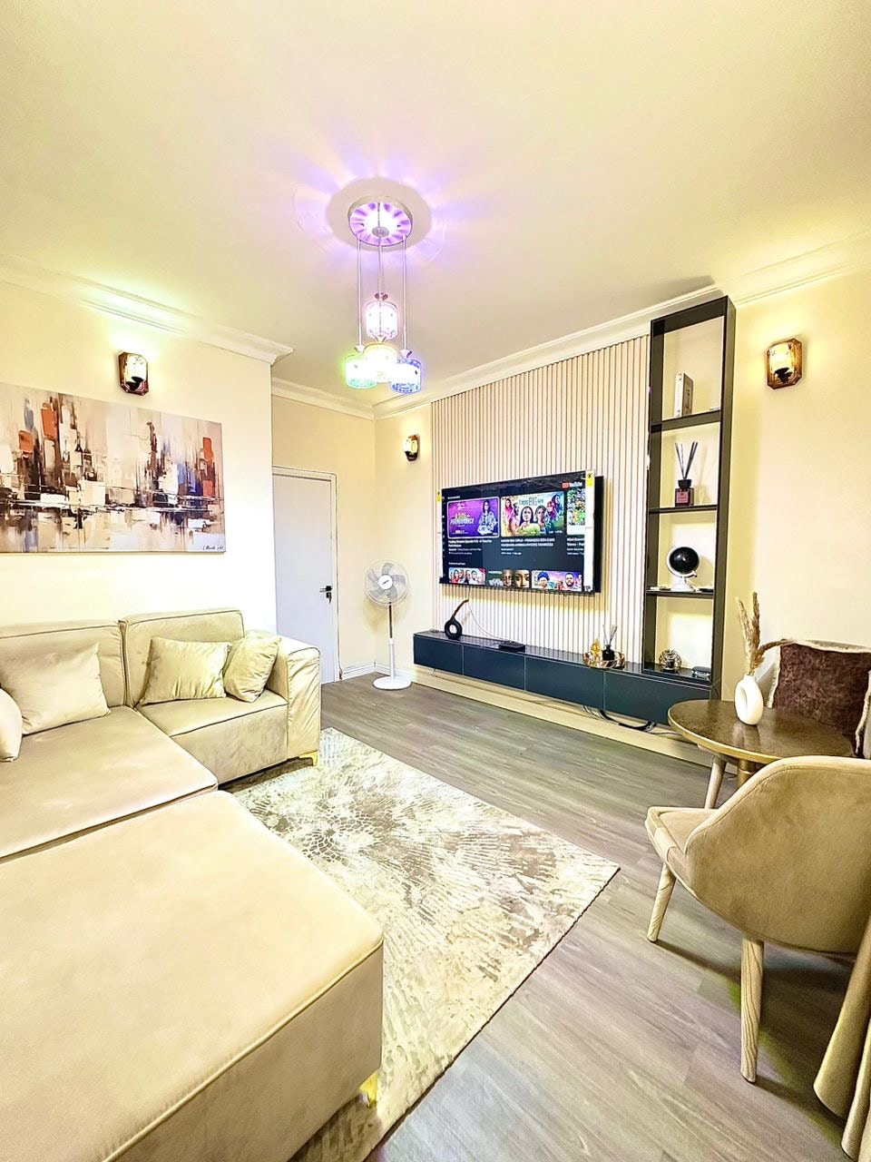 A modern living area features a soft beige sectional sofa paired with a stylish chair. A flat-screen TV is mounted on a decorative wall unit, while a fan stands nearby. Ambient lighting adds a welcoming feel, and an abstract painting adorns the wall.
