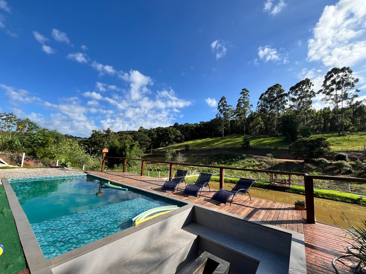 An outdoor infinity pool reflects a clear blue sky, surrounded by a wooden deck with several lounge chairs positioned for relaxation. Lush green hills serve as a backdrop, providing a natural and serene setting.