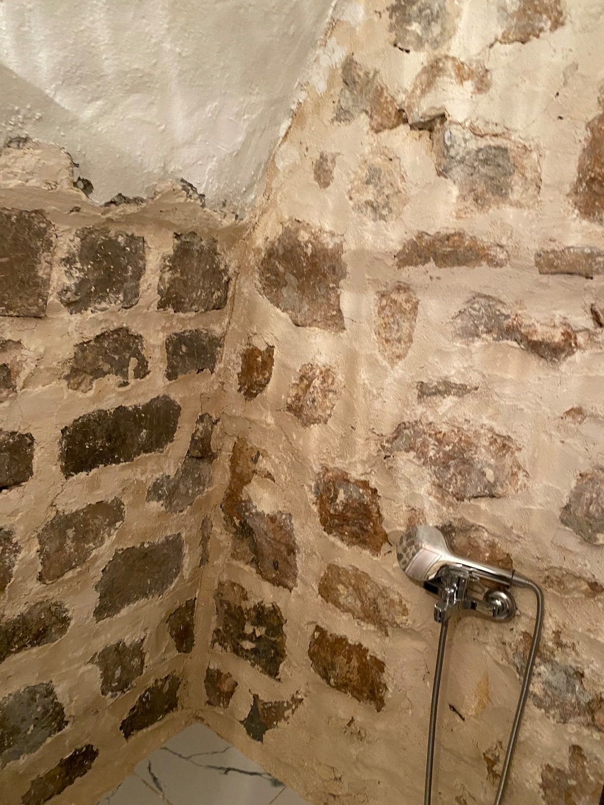 The image captures a corner of a shower area featuring rustic stone walls, revealing a blend of rough and smooth textures. A shower fixture is mounted on the wall, complementing the natural stone aesthetic. Natural light creates subtle shadows, enhancing the stone's features.