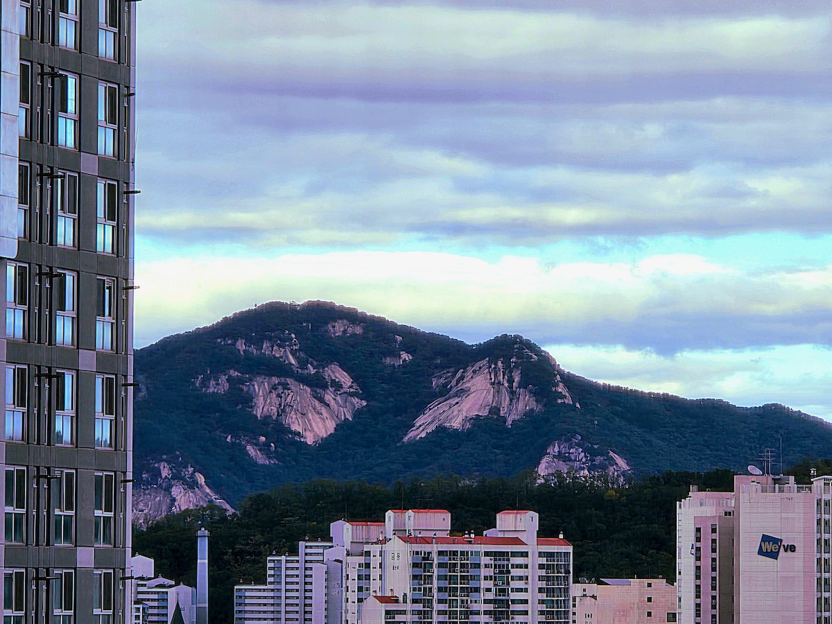 A distant view of Bukhan Mountain is observed, showcasing its rocky peaks and green slopes under a softly clouded sky. The mountain is framed by modern buildings in the foreground, highlighting the contrast between nature and urban life.