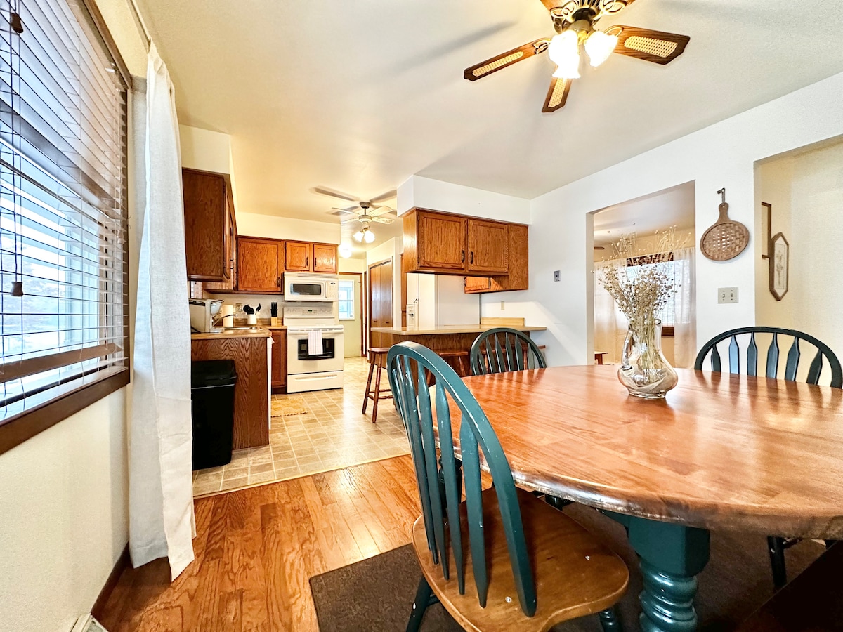 The dining area features a round wooden table surrounded by blue-green chairs, illuminated by a ceiling fan. A warm, inviting kitchen is visible in the background, with oak cabinetry and modern appliances. Natural light enters through windows dressed with blinds.