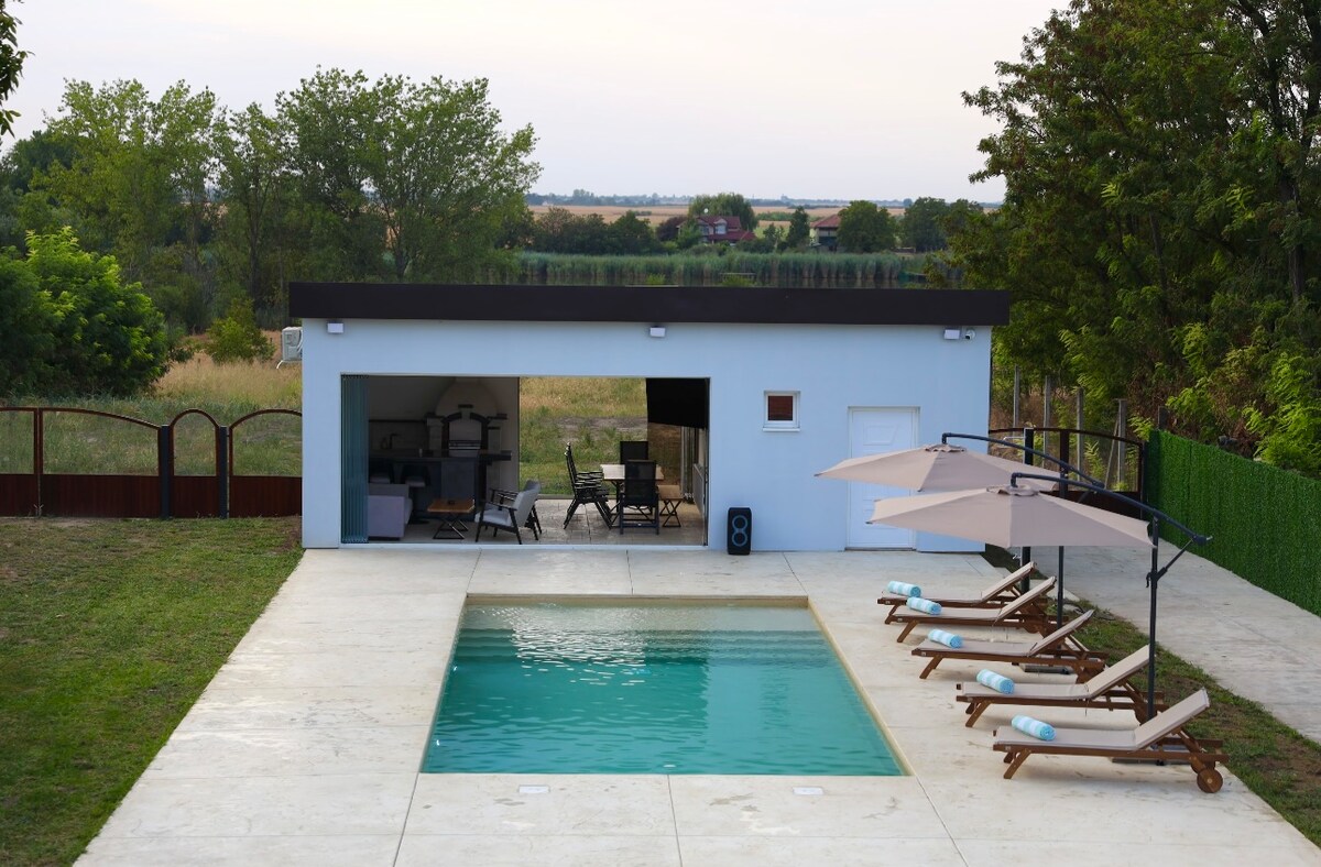 An outdoor space features a serene pool surrounded by lounge chairs. A small building with large windows complements the setting. There are shaded areas provided by umbrellas, and the landscape includes lush greenery and an unobstructed view of the distant fields.