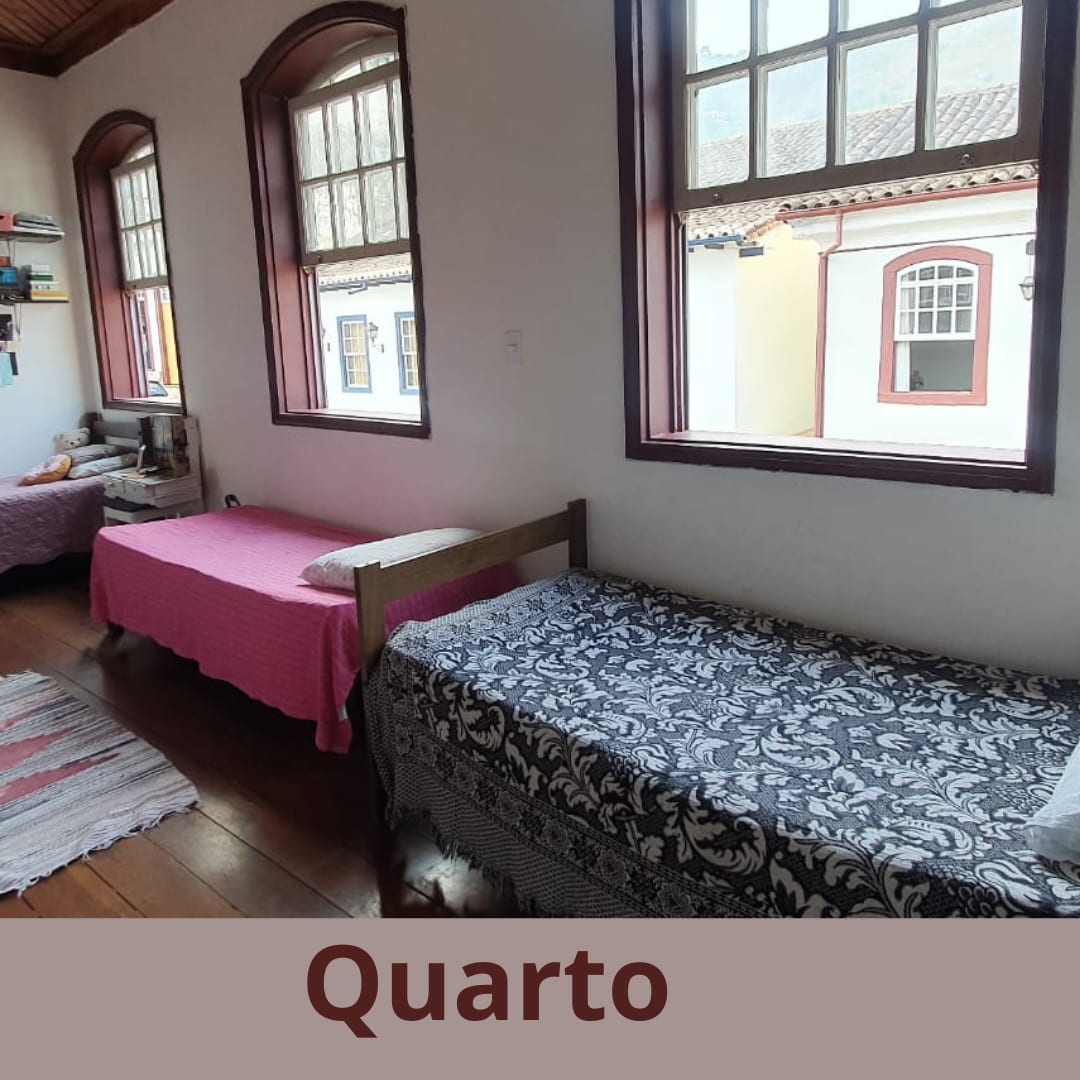 A bright room features two single beds, one adorned with a pink coverlet and the other with a patterned blanket. Large windows allow natural light to fill the space, framed by wooden trim, while a colorful rug adds texture to the wooden floor.
