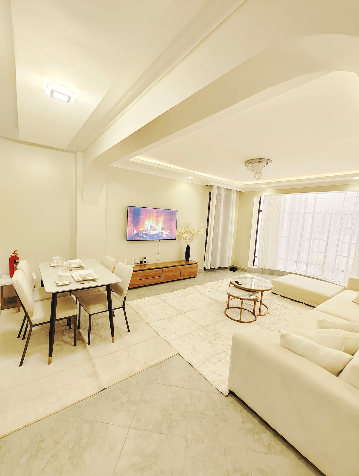 The open-plan living area is equipped with a modern smart TV mounted on the wall. A cozy seating arrangement surrounds a light-colored coffee table. A well-set dining table for four is positioned near a large window, allowing natural light to enhance the space.