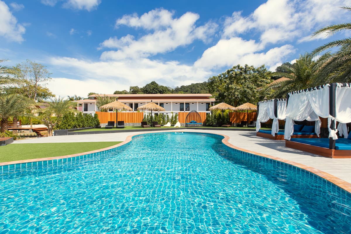 A large swimming pool with clear blue water is surrounded by palm trees and vibrant greenery. Poolside cabanas offer shaded seating areas, while the backdrop features a series of low-rise buildings with orange accents. A bright sky with scattered clouds enhances the scene.