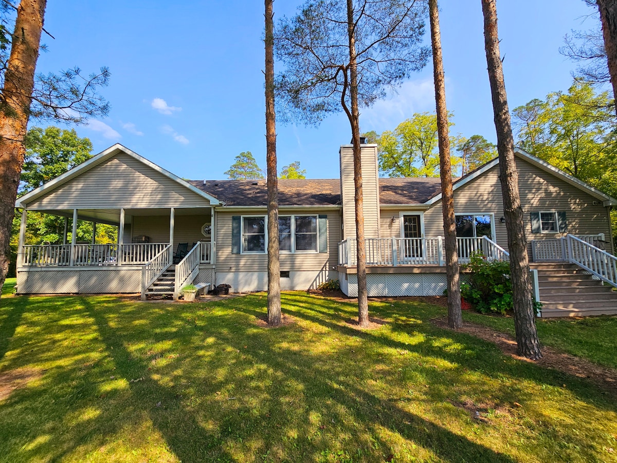 A single-story home is framed by tall trees, set against a clear blue sky. The exterior features a large porch with railings and multiple steps leading up to the entrance. Lush green grass surrounds the house, enhancing the serene atmosphere.