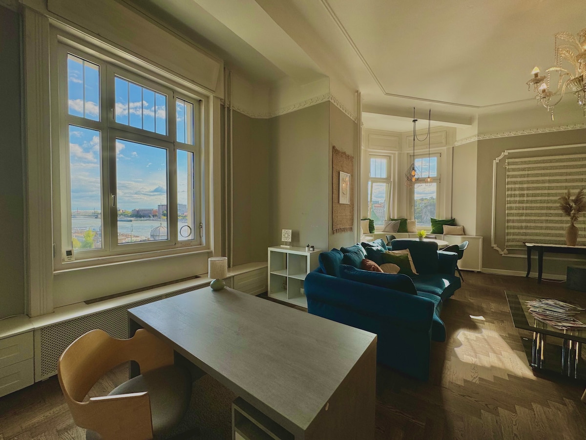 A spacious living area features a large window that frames scenic views of the outside. Designer furnishings include a modern blue sofa and a sleek wooden desk, allowing for a comfortable and functional space. Natural light illuminates the room, highlighting its elegant decor.