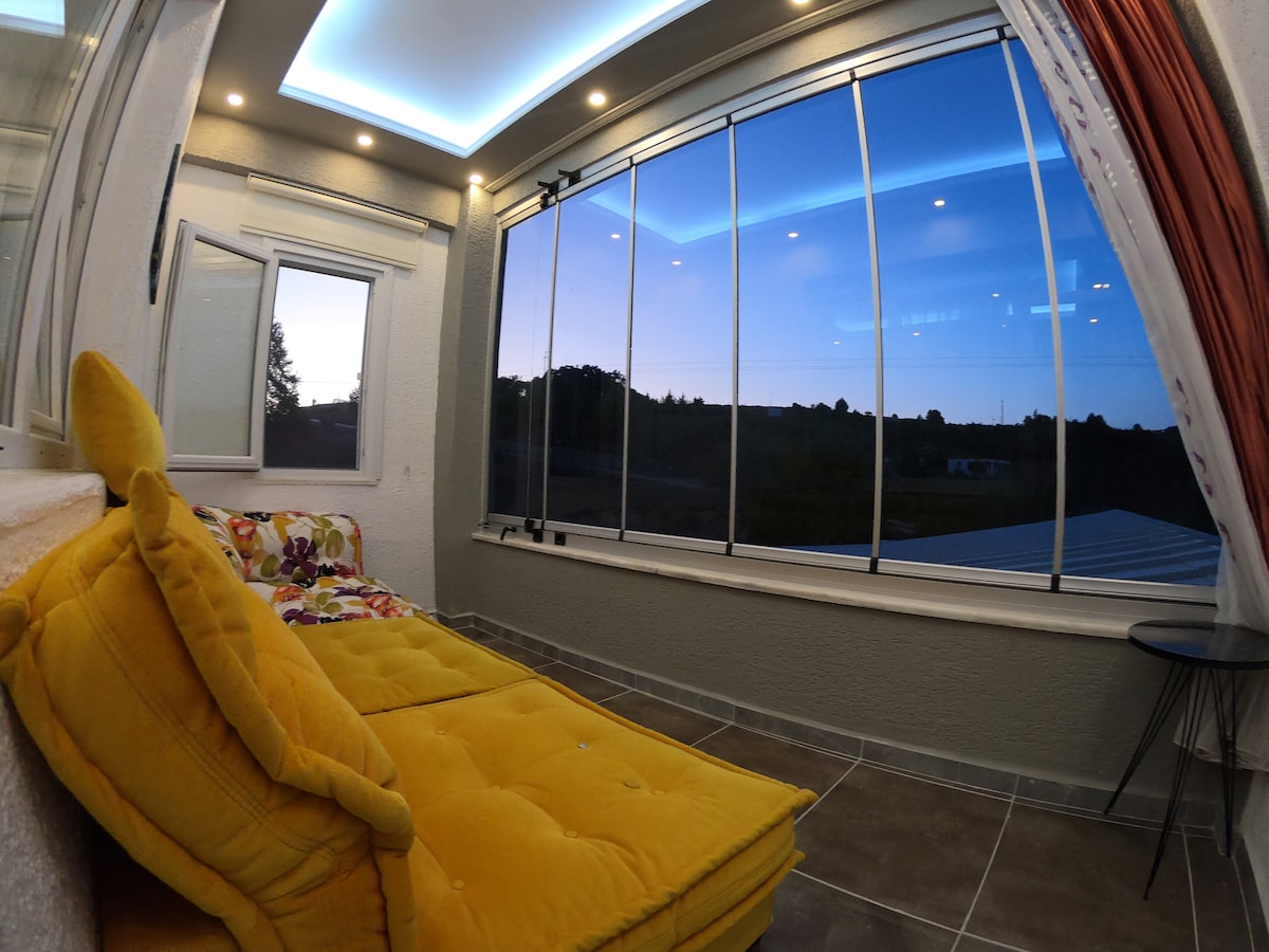 A cozy seating area includes a bright yellow sofa adorned with colorful cushions. Large glass windows provide an expansive view of the outdoors, illuminated by soft lighting overhead. The room features a combination of warm tones and modern design elements.