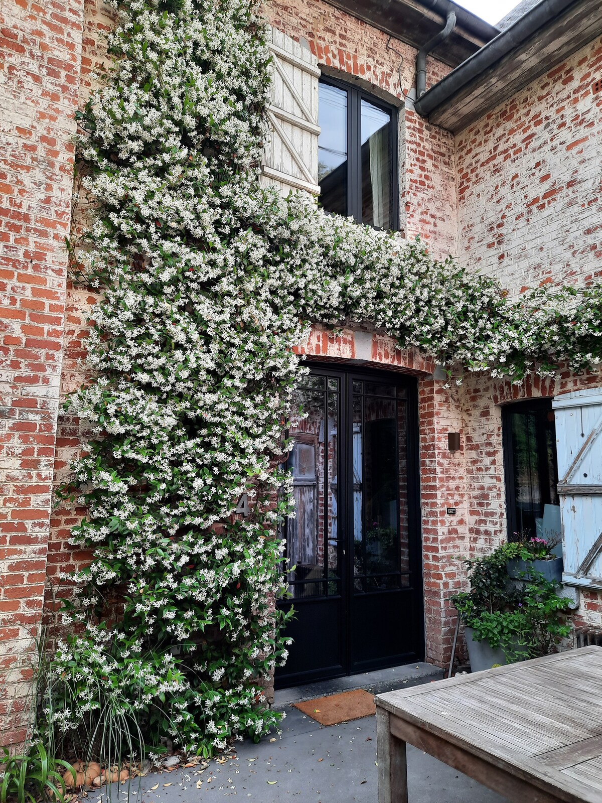 A rustic brick exterior features a striking floral vine cascading over the entrance, enhancing the inviting facade. Large glass doors provide a glimpse into the interior, while weathered shutters and a wooden table nearby add to the charming outdoor setting.