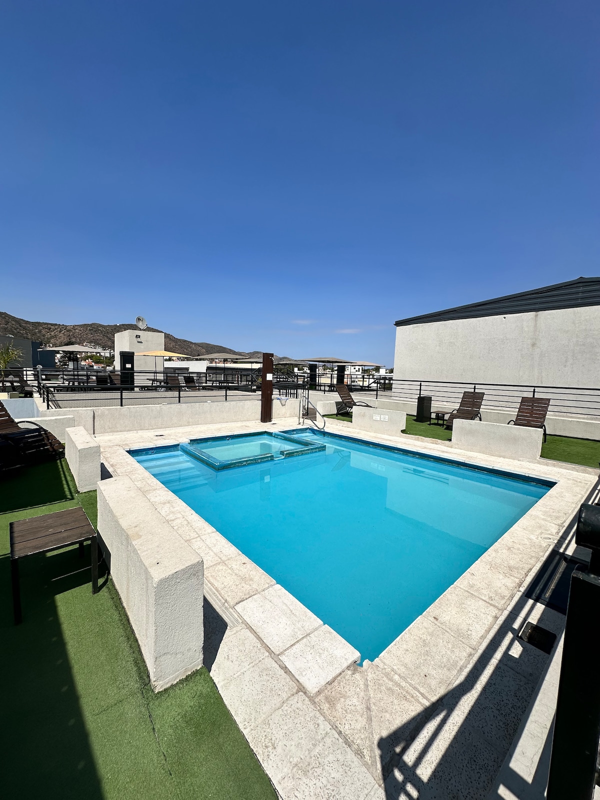 An outdoor pool is set on a rooftop, surrounded by a smooth stone deck. Several lounge chairs are arranged along the perimeter, with a scenic view of distant mountains and blue skies above. The pool features a built-in hot tub area.