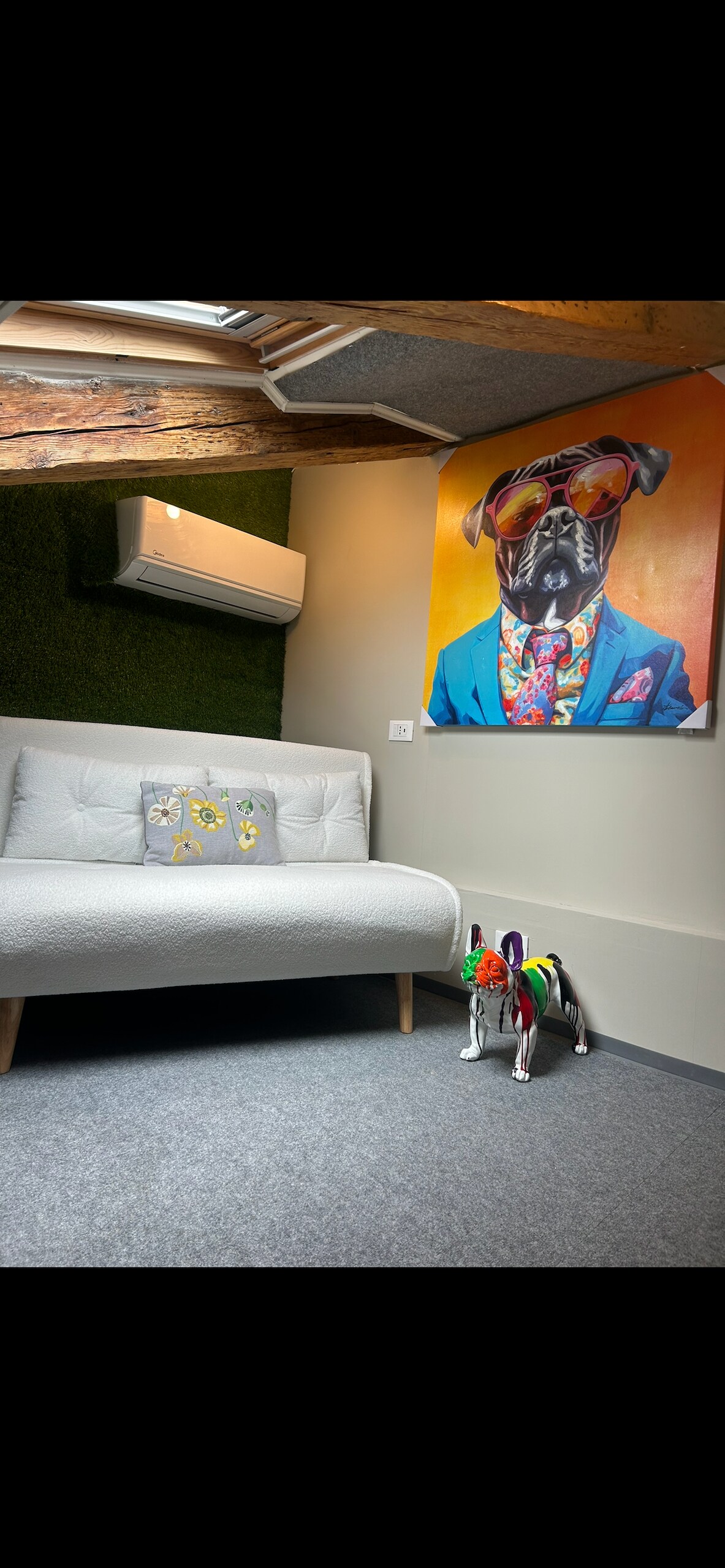 A cozy corner features a light-colored sofa and a colorful dog-themed artwork on the wall. A decorative dog figure and a small accent pillow with floral design are present. Exposed beams and air conditioning unit are visible, enhancing the inviting atmosphere.
