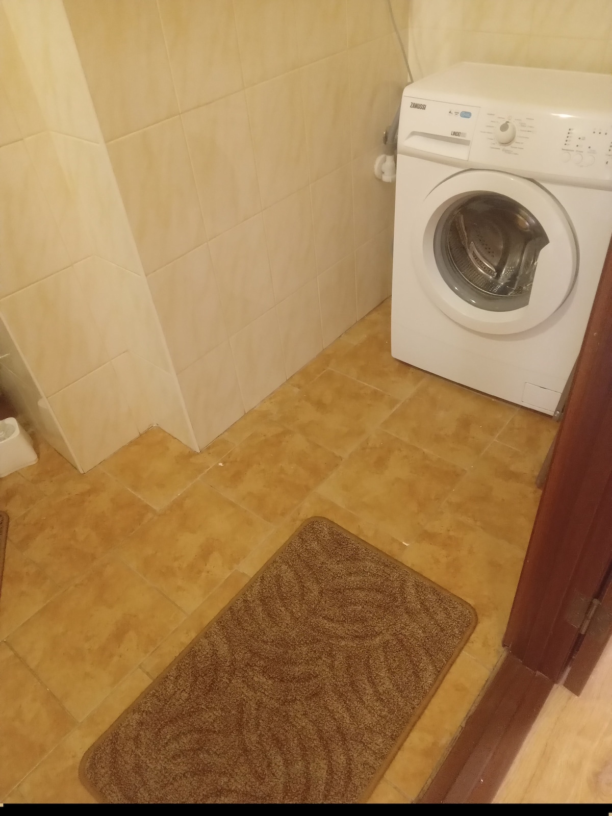 A laundry area is presented with a washing machine positioned against a tiled wall. The floor is covered with light brown tiles, complemented by a textured mat in front of the machine. The space appears clean and functional, with neutral colors enhancing its practicality.