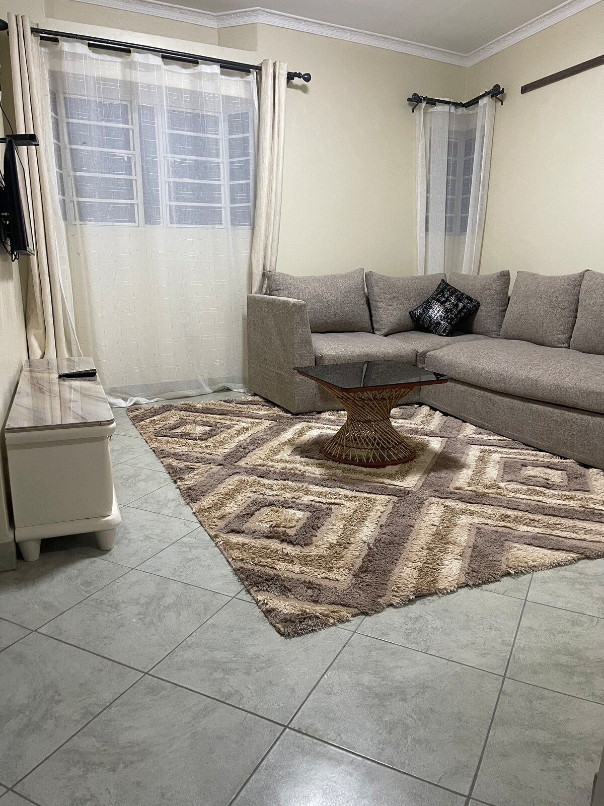 A cozy living area features a corner sofa complemented by soft pillows. A patterned area rug adds texture to the tiled floor, while light drapes frame the window, allowing natural light to fill the space. A small, round table sits in front of the sofa.