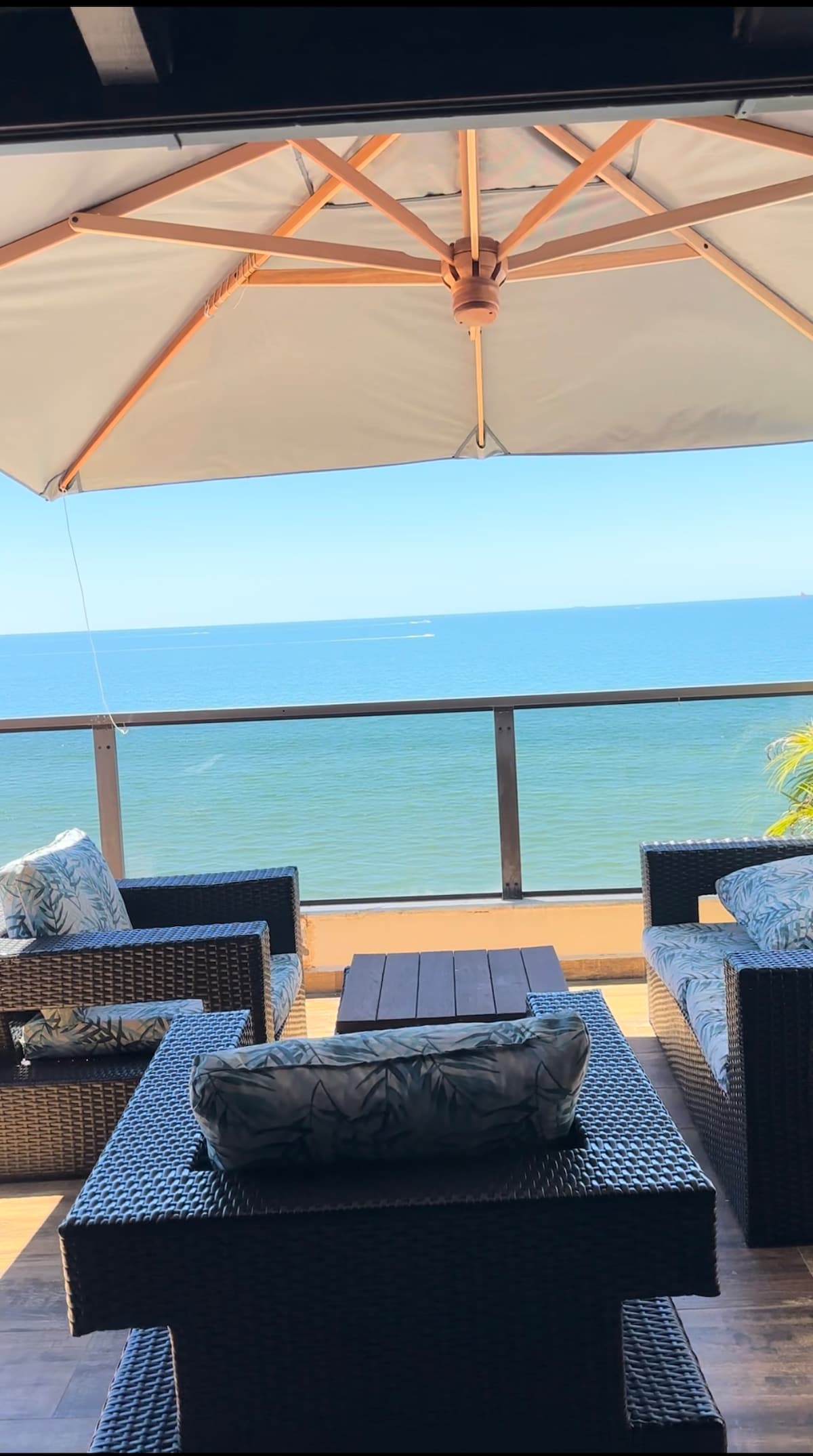 A seating area is presented with woven furniture, featuring two cushioned chairs and a coffee table. A large umbrella provides shade while overlooking a vast ocean view. The tranquil colors of the sea blend seamlessly with the clear sky.