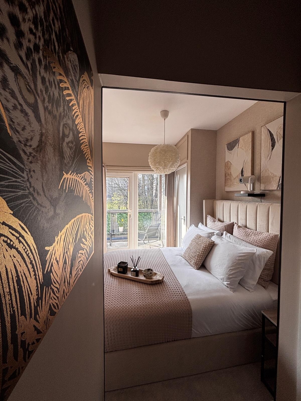 A cozy bedroom is framed by soft neutral tones, featuring a spacious bed adorned with multiple pillows. A warm artwork of a leopard is displayed on the wall. Natural light enters from the balcony, creating a bright and serene atmosphere.