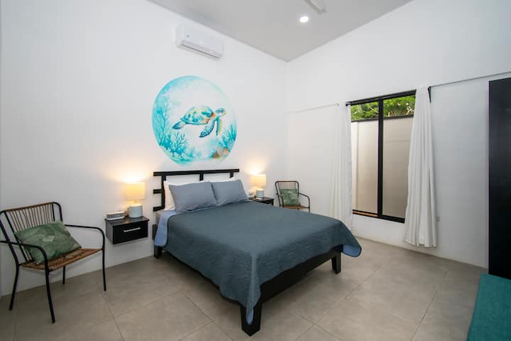 Guest bedroom at Coral Dream House featuring a queen-size bed, a/c, two decorative chairs, a comfortable bench and a spacious closet, offering a cozy and serene retreat within the home.