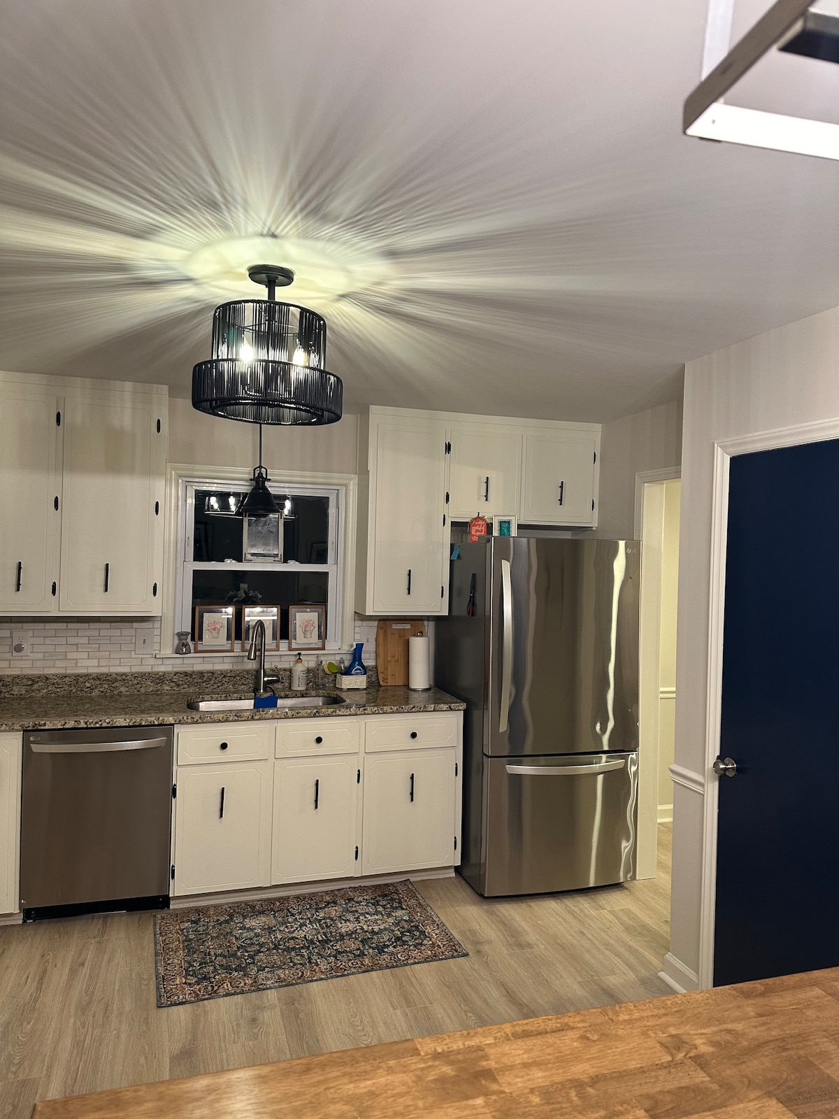 The kitchen features modern appliances, including a stainless steel refrigerator and a dishwasher. White cabinetry lines the walls, complemented by a patterned rug on the floor. A pendant light casts a soft glow over the workspace, enhancing the functionality of the area.
