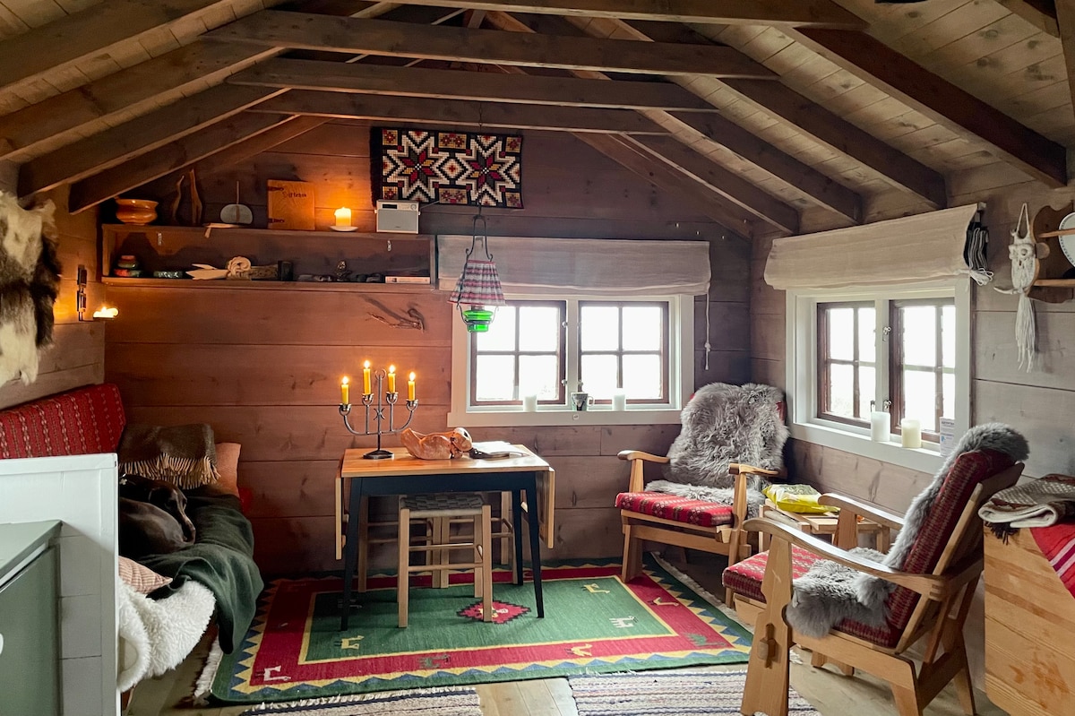 The interior of a rustic cabin features a wooden ceiling with exposed beams. A dining table is surrounded by two wooden chairs, with a cozy seating area that includes a plush blanket draped over a bench and another chair. Candles and a soft rug add warmth to the space.