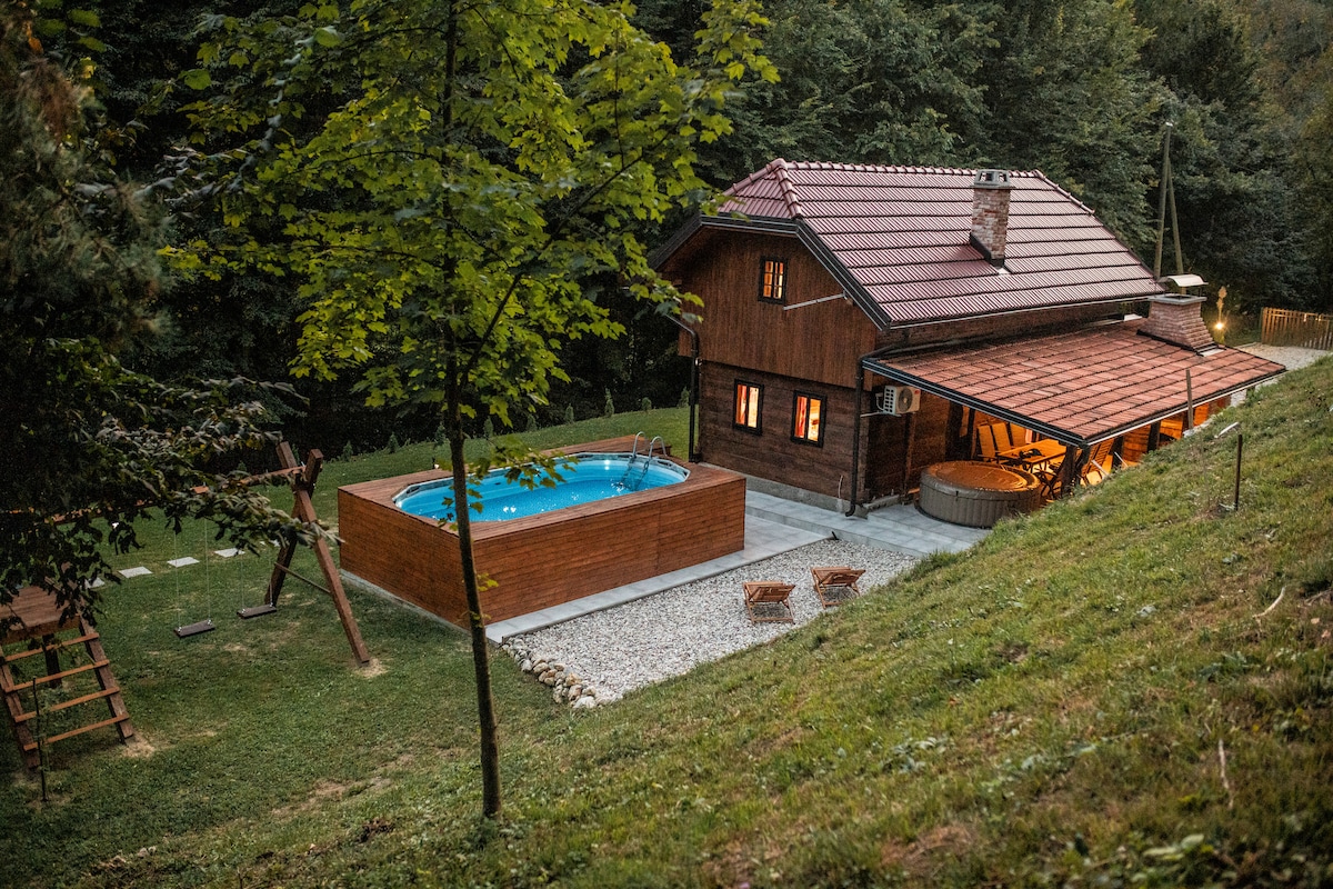 A charming wooden house is nestled among trees, featuring a private pool and hot tub in the outdoor area. The soft glow from the windows creates a warm ambiance. A grassy yard includes a playground and seating area, providing space for relaxation and play.