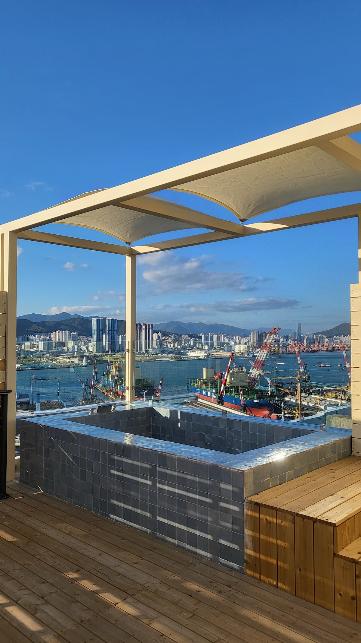 A rooftop terrace showcases a jacuzzi surrounded by wooden decking. The panoramic view features a bustling harbor with boats and city buildings, beneath a clear blue sky. The structure is partially shaded by a modern canopy, providing a private outdoor space.