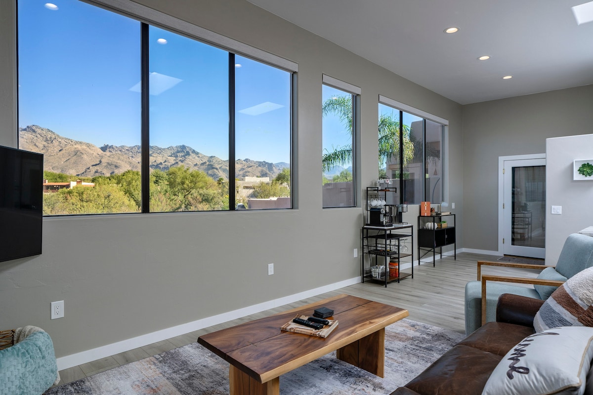A bright and airy living space features large windows with mountain views, allowing natural light to illuminate the room. A wooden coffee table sits center stage, complemented by comfortable seating options. The open layout enhances the connection to the outdoors, showcasing a tranquil setting.