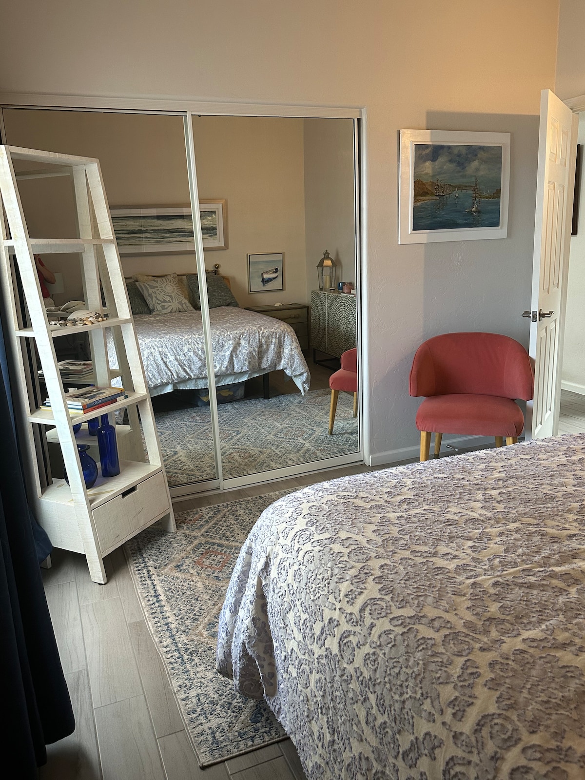 A cozy bedroom is showcased with a spacious bed draped in a patterned bedspread. A mirror closet reflects the room, adding depth. A decorative chair sits nearby with a small side table, while art is displayed on the wall, enhancing the serene ambiance.
