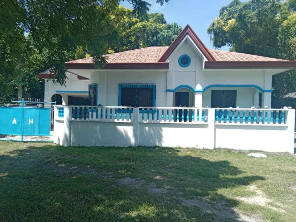 The exterior of the cozy rest house is shown, featuring a gated entrance with a turquoise gate. The house has a simple design with a sloped roof and large windows. Surrounding grass is visible, providing a natural setting under the shade of trees.