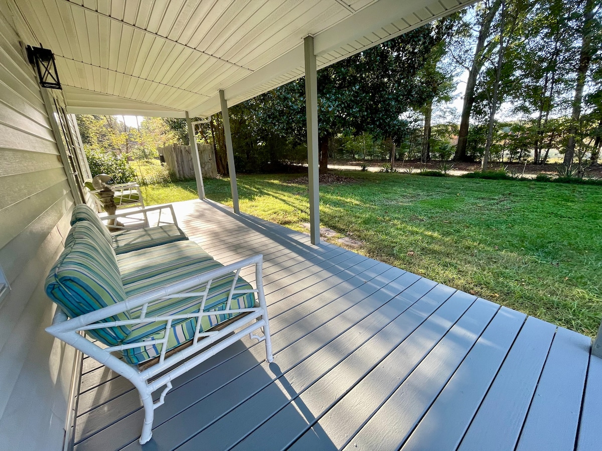 A spacious covered porch features a striped sofa, inviting relaxation. The floor is painted in a light gray, and sunlight filters through the trees, illuminating the green grass and surrounding landscape. The peaceful outdoor area offers a serene setting for enjoying nature.