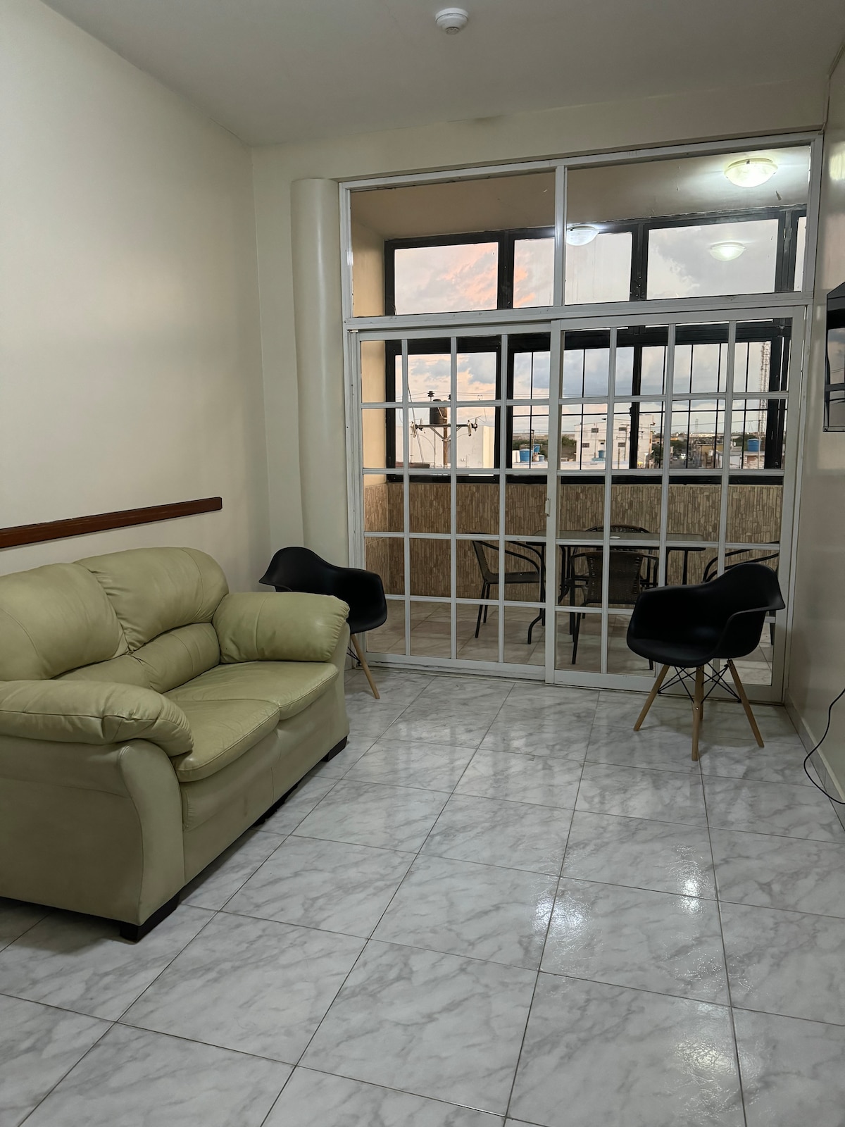 A well-lit seating area features a light-colored couch and two black chairs. Large glass doors lead to a balcony, offering views of the surroundings. The floor is tiled in glossy white, enhancing the spacious feel of the room.