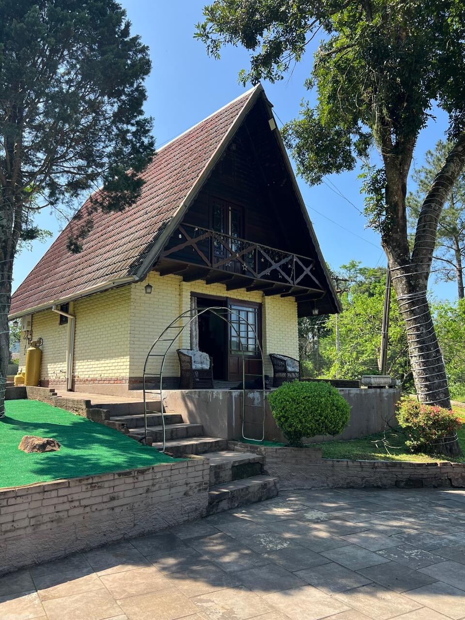 A charming A-frame house is positioned among lush greenery, featuring a brick walkway leading to the entrance. A shaded balcony is visible above, complemented by wooden railings. The well-maintained exterior showcases light-colored brick, inviting trees, and manicured landscaping.