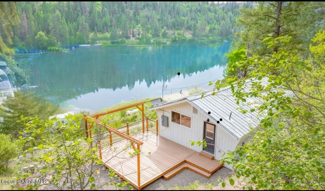 A charming lake house is shown from an elevated angle, surrounded by lush greenery. A spacious wooden deck offers views of the lake, while the house features a light-colored facade with large windows. The tranquil water reflects the surrounding mountains and trees.