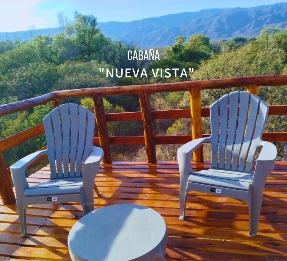 Two gray adirondack chairs are positioned on a wooden deck, creating a serene spot for relaxation. A small round table enhances the inviting space, with a backdrop of lush greenery and mountains visible in the distance.