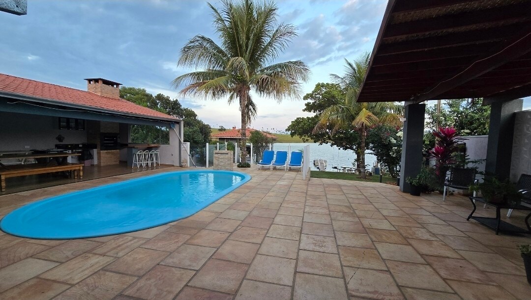 A serene outdoor area features a crystal-clear pool surrounded by a spacious stone patio. Sun loungers are arranged nearby, complemented by palm trees and lush greenery. The view extends to a calm body of water, enhancing the relaxing ambiance of the space.