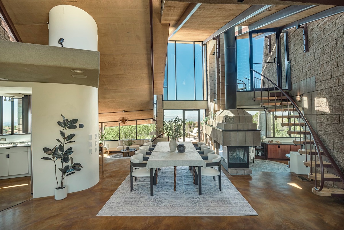 An open-concept living area features high ceilings and generous natural light from large floor-to-ceiling windows. A central dining table is surrounded by comfortable chairs, while a stylish staircase leads to upper levels. A unique fireplace adds a focal point to the space.