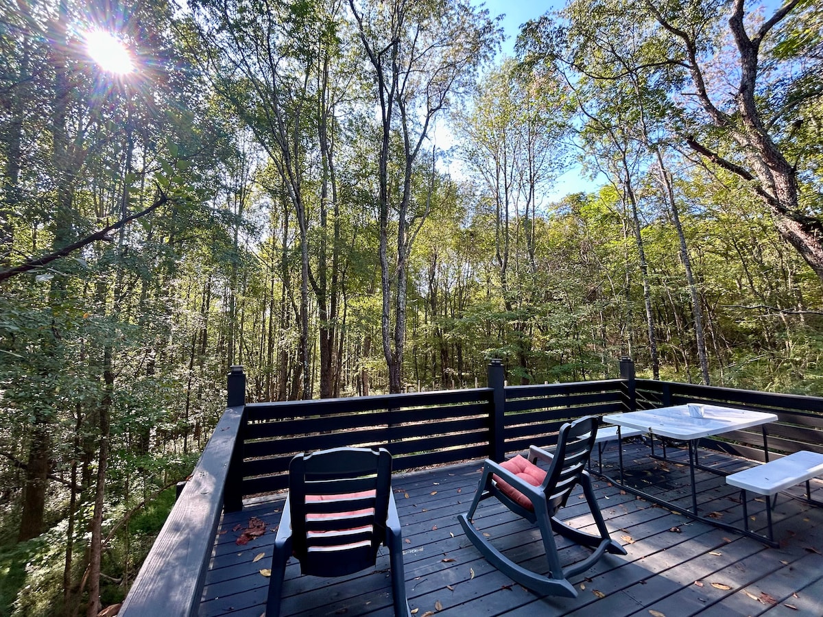 A spacious deck is surrounded by lush trees, providing a serene wooded view. Two rocking chairs are positioned for relaxation, while a table offers space for dining or games. Soft sunlight filters through the leaves, creating a peaceful atmosphere.
