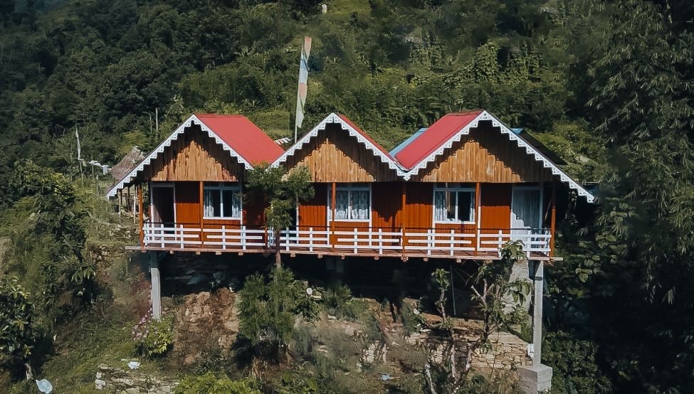 Two wooden cabins with red roofs are positioned on a hillside, surrounded by lush greenery. Large windows are visible, offering scenic views. The cabins are elevated on sturdy pillars, providing a sense of openness and connection to the natural environment.