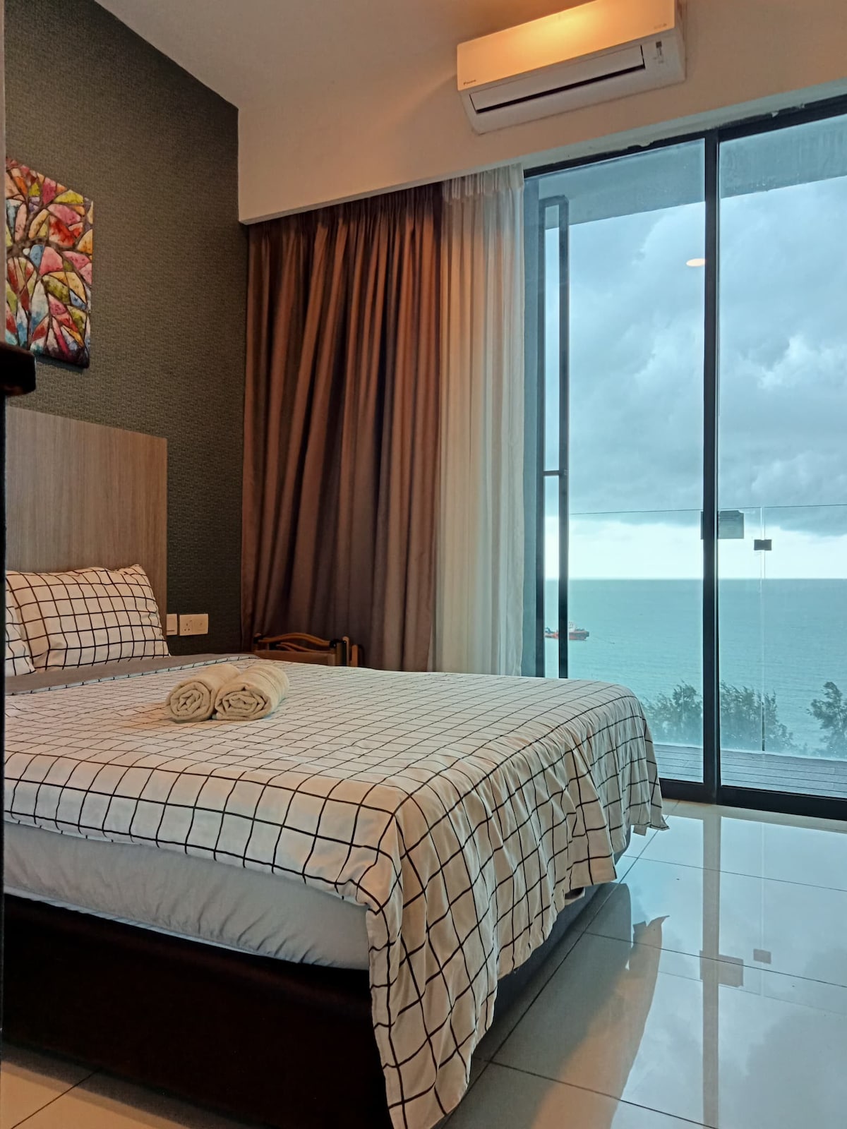 A bright sleeping area features a spacious bed dressed in a checkered bedspread. Floor-to-ceiling windows provide views of the seafront, with sheer curtains allowing soft natural light to filter in. Towels are neatly arranged on the bed, enhancing the serene ambiance.