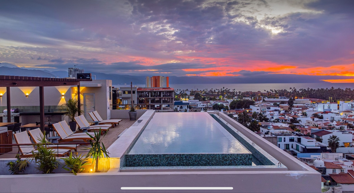 A modern rooftop infinity pool extends toward a colorful sunset, offering panoramic views of the surrounding city and ocean. Lounge chairs are strategically placed along the edge, framed by greenery, creating a serene space for relaxation.