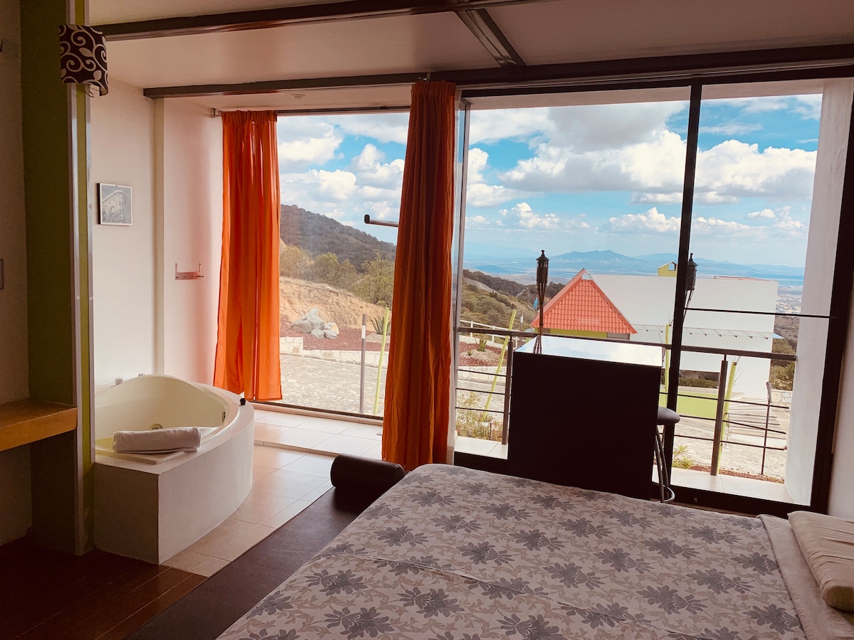 A spacious room features a king-size bed and a modern Jacuzzi tub positioned next to a large window. Soft orange curtains frame the view of the valley and distant mountains. Natural light fills the area, enhancing the room's inviting ambiance.
