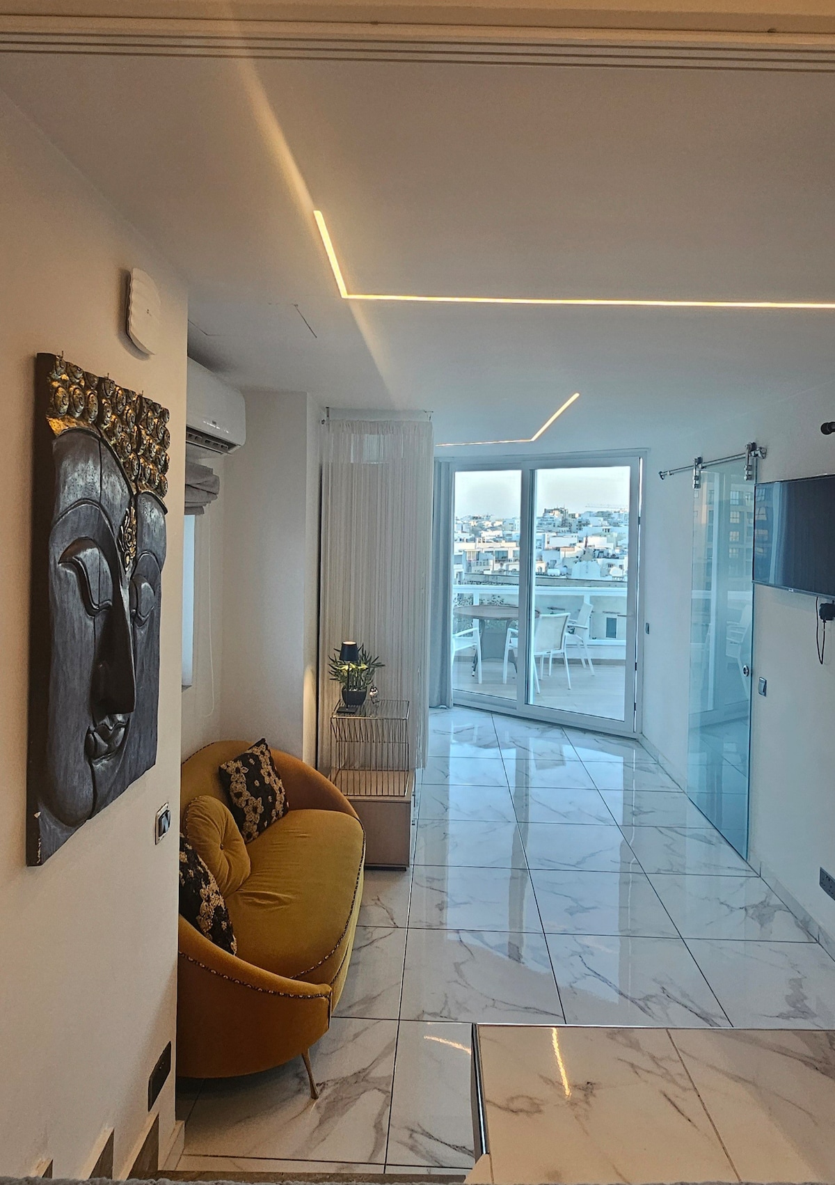 A modern hallway features a striking artwork of a face on the wall. A yellow couch is positioned beside a glass door leading to a balcony. Natural light filters through sheer curtains, reflecting off the glossy marble floor, and creating an open and airy feel.