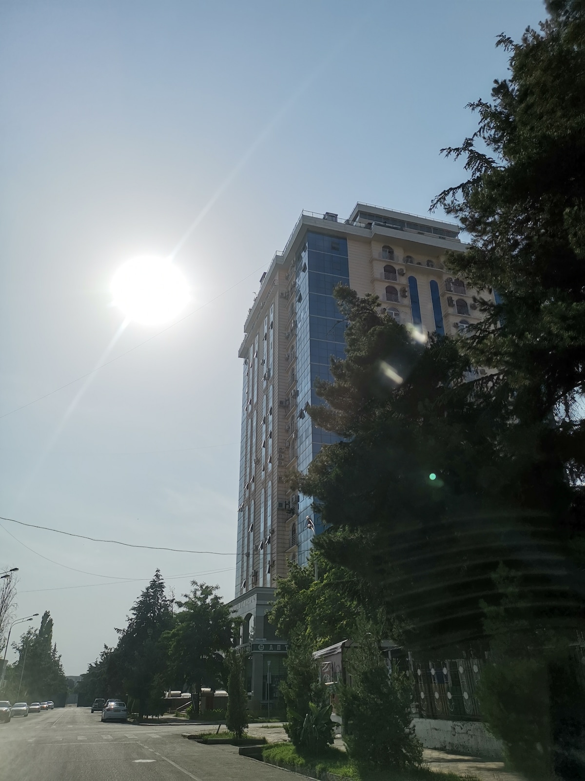 A modern high-rise building is observed, featuring a glass facade that reflects sunlight. Lush greenery is present around the base, and a clear sky with the sun shining above adds brightness to the scene.