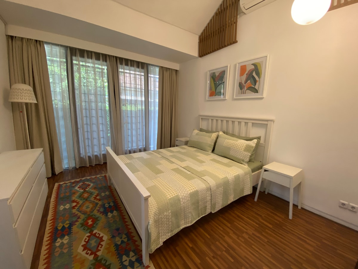 A bright sleeping area features a spacious bed with soft green linens and decorative pillows. Two light-colored artworks are displayed above the bed. Natural light filters through sheer curtains, illuminating the room's wooden flooring and a colorful area rug.