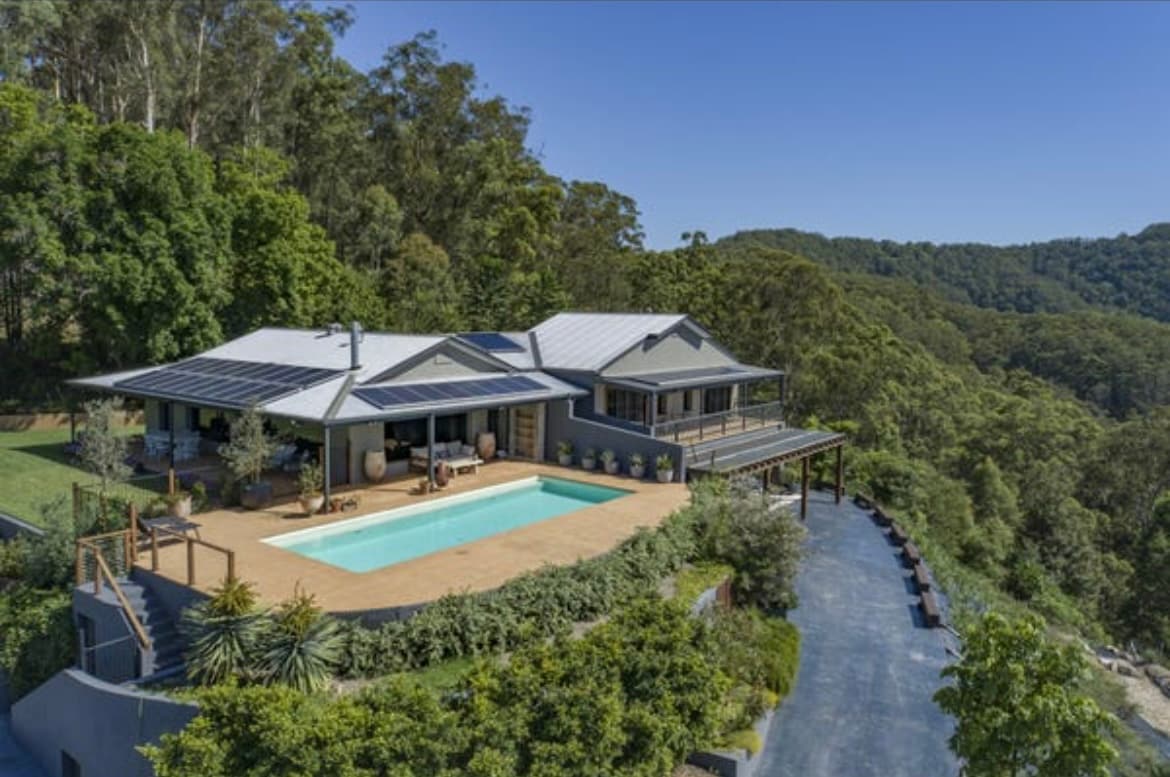 A modern, hillside home is surrounded by lush greenery, featuring a spacious deck with a resort-style pool. Solar panels adorn the roof, reflecting an eco-friendly design. Terraced landscaping creates a harmonious blend with the natural environment, offering scenic views of the surrounding bushland.