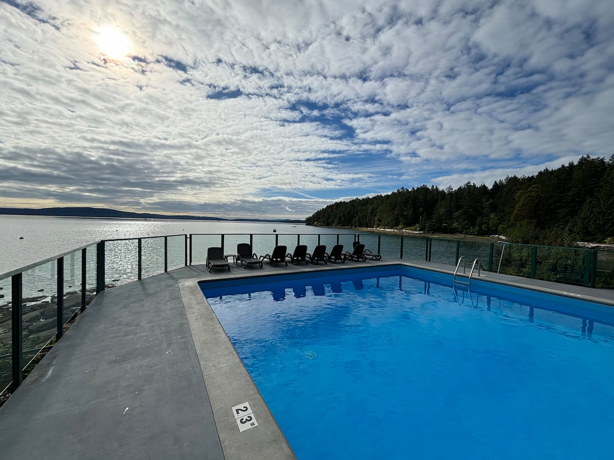 A clear blue swimming pool is positioned overlooking the water, surrounded by a glass railing. Several black lounge chairs are arranged along the pool's edge, while trees line the shoreline in the background beneath a partially cloudy sky.
