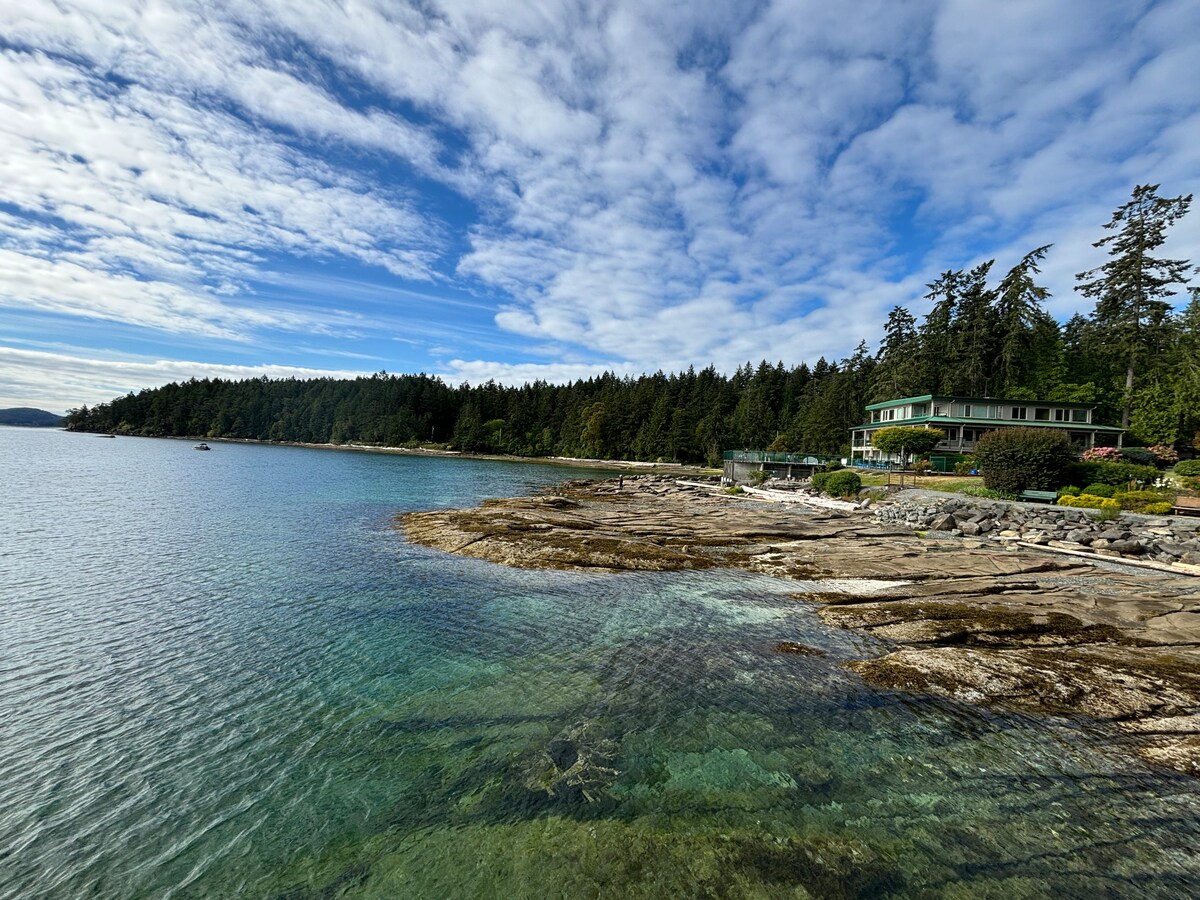 The waterfront landscape features clear blue waters gently lapping against rocky shores. Lush green trees line the backdrop, while a coastal building is seen in the distance. The sky is partly cloudy, contributing to a serene and inviting atmosphere.