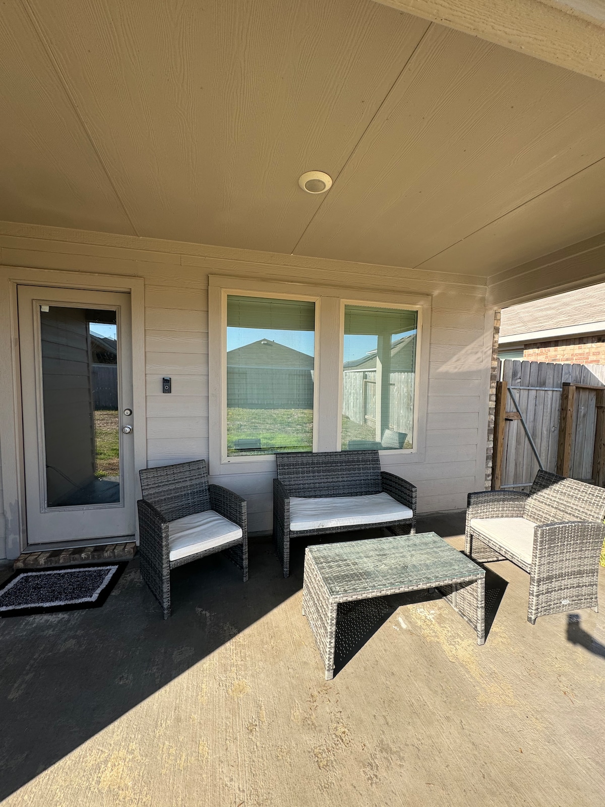 An outdoor seating area features a small table surrounded by four wicker chairs, two of which are cushioned. Large windows provide views of the surrounding outdoor space. The entrance door is visible, leading to the interior.