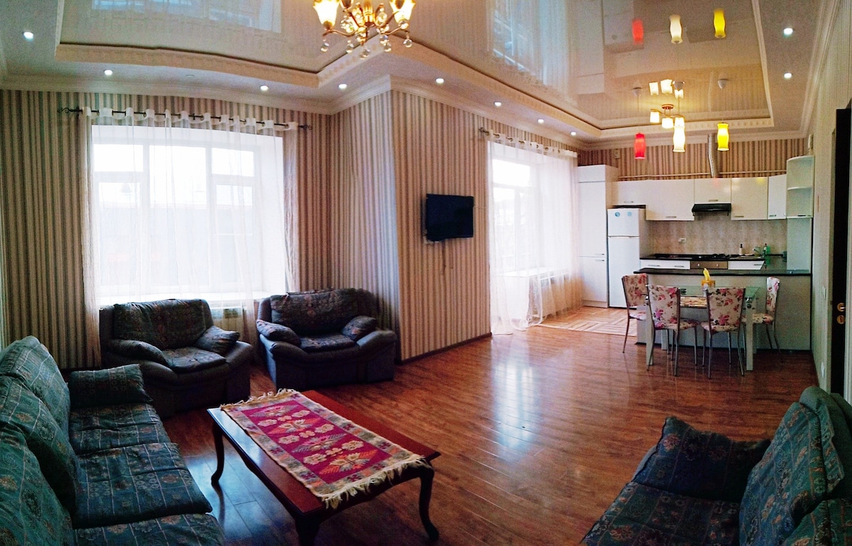 A spacious living area is equipped with comfortable seating arrangements, featuring plush sofas and a coffee table. The dining area accommodates several chairs around a table. Large windows allow natural light to fill the room, and decorative lighting adds a touch of color to the ceiling.