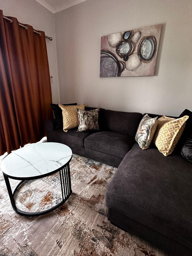 A dark sectional sofa is complemented by decorative pillows in various patterns. A round coffee table with a marble top sits on a plush area rug featuring warm tones. Natural light filters through the brown curtains, enhancing the cozy indoor space.